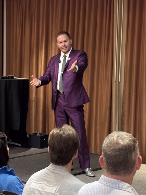 Michael, in a purple suit, speaks to attendees at a confrence.